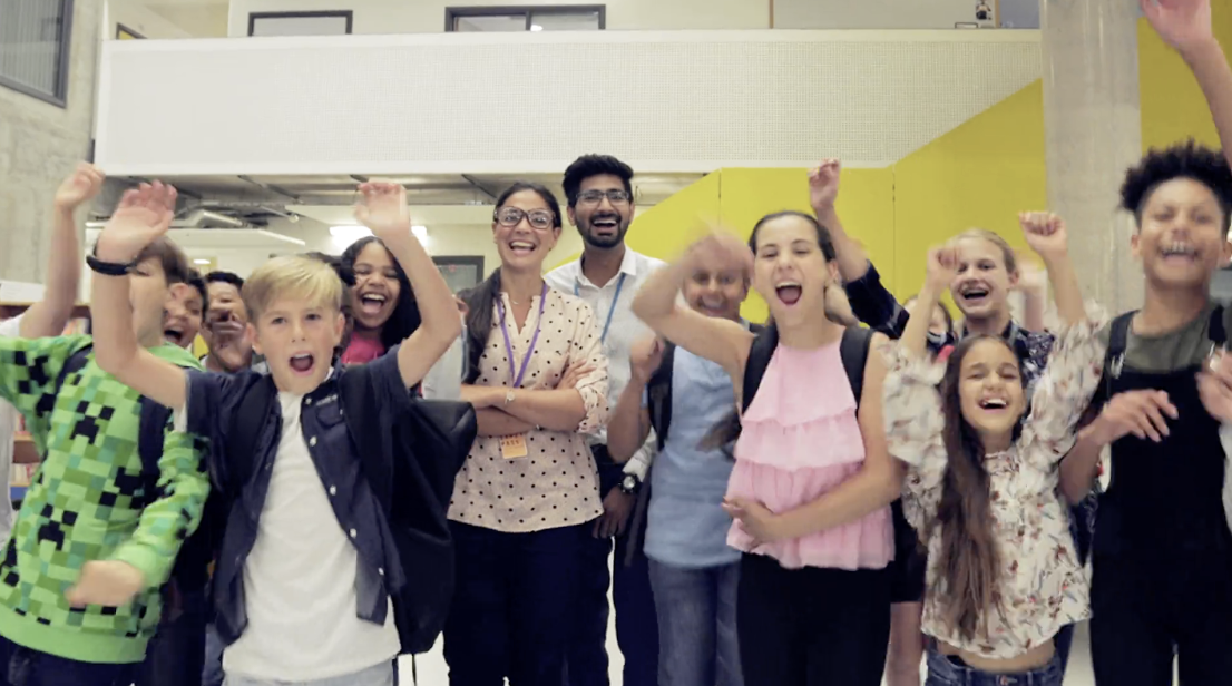 Photo of two teachers in the school hallway with a bunch of joyful children jumping up and down