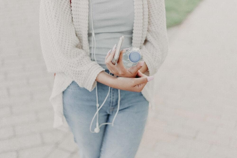 Woman wearing white carigain and light jeans carrying smart phone with headphones and single-use water bottle close to body