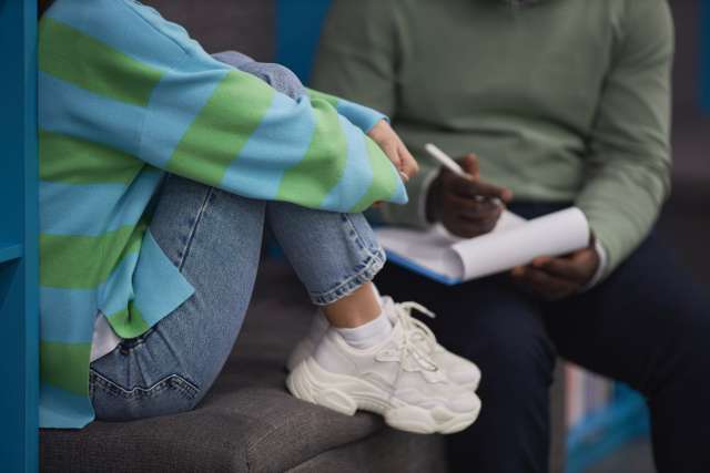 A therapist talks with a teenage girl who is sitting up with her knees curled to her chest.