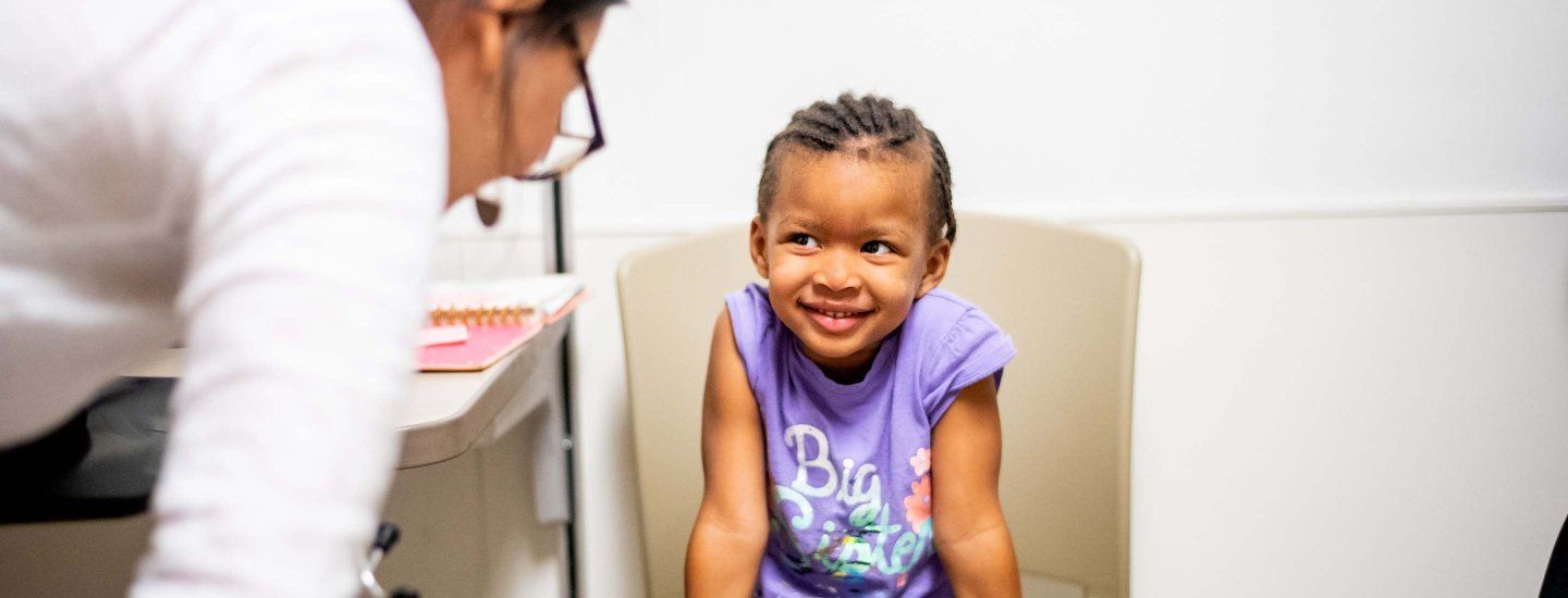 A child smiles shyly as a clinician leans down to speak to her