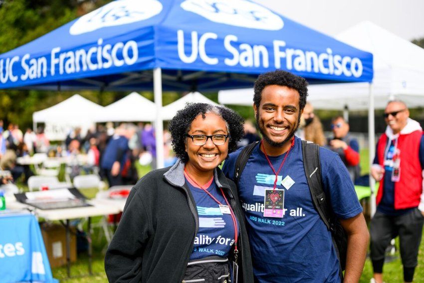 A faculty member and a staff research associate wear matching AIDS Walk shirts in front of a tent that says UC San Francisco.