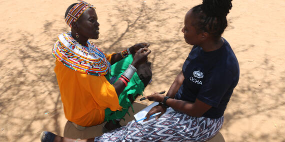 Two women sit on bare ground talking to each other.