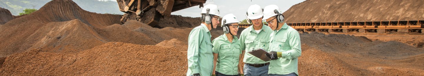 Foto de 3 homens e uma mulher em uma área de operação olhando para a prancheta que um deles está segurando. Todos estão usando uniforme da Vale.