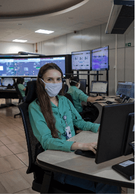 Photo of a woman sitting on a chair working on a computer in an office with several monitors and screens in the background. She has long dark hair and is wearing a uniform, a green button-up shirt, badge, and face mask.