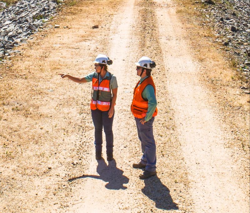 A man and a woman – facing each other – in an operational space. The two wear light green shirts, goggles, ear muffs, and white helmets.