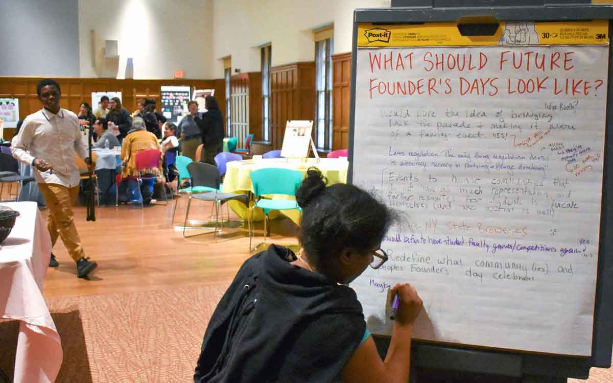 A very large room with wood-paneled walls and windows. An event is taking place here. In the foreground, a person with glasses and black hair in two buns is writing on a whiteboard, they’re back to the viewer. The whiteboard has a handwritten title at the top reading “What should future founder’s days look like?” Below it are written suggestions in different handwriting. In the background, a group of people sit at a table.