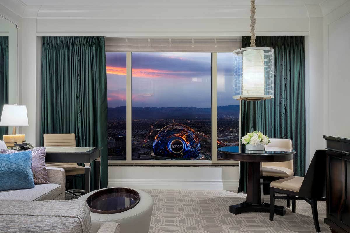 Hotel room interior with modern furniture and a view of a cityscape at sunset through a large window.