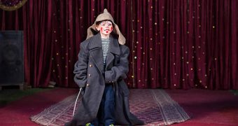 Talented boy standing and holding a sword while wearing oversized retro gray coat and Russian hat over modern casual clothes during theatrical play on stage