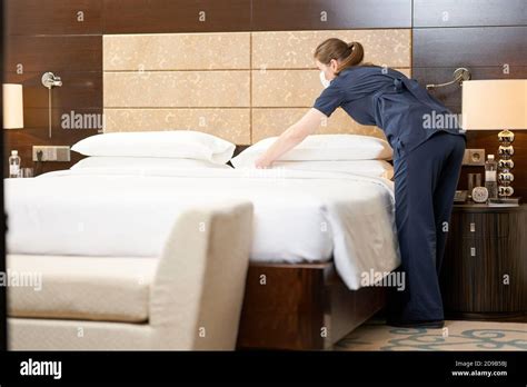 Full length photo of maid in mask putting clean pillows on the bed in the hotel room Hotel ...