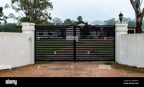 Black modern metal driveway property entrance gates set in concrete fence, lights, trees Stock ...