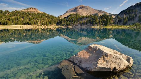 a large rock sitting in the middle of a lake surrounded by trees and mountains on a sunny day