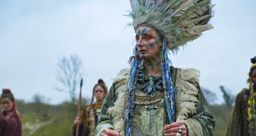 photo of a screenshot from the tv show Brittania. It features an elderly woman wearing a bluish white feathered headdress extremely reminiscent of Native American war bonnets. She is also wearing green jewelry and a feathered necklace. Characters in the background, while blurred, also have similar clothing reminiscent of Native American traditional clothing