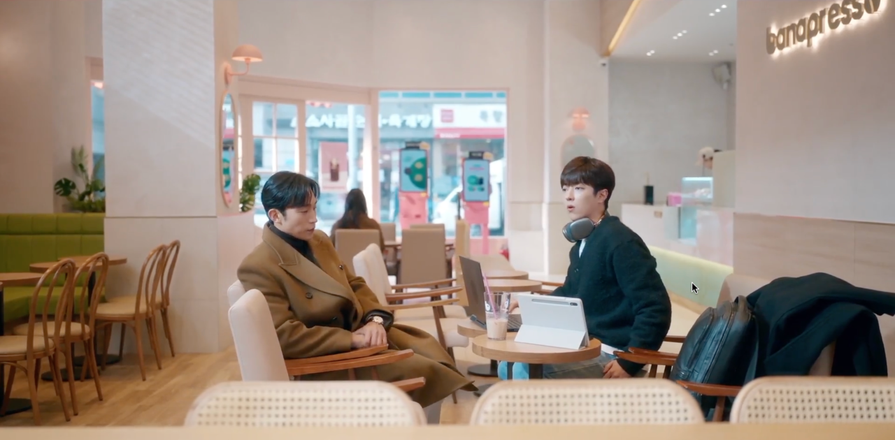 another screenshot from "no gain no love". it's from the same scene, but zoomed out so you can that we're at a cafe. hajun's sitting at a round table in the cafe working on his laptop, with headphones on. his boss sits across from him dejectedly.