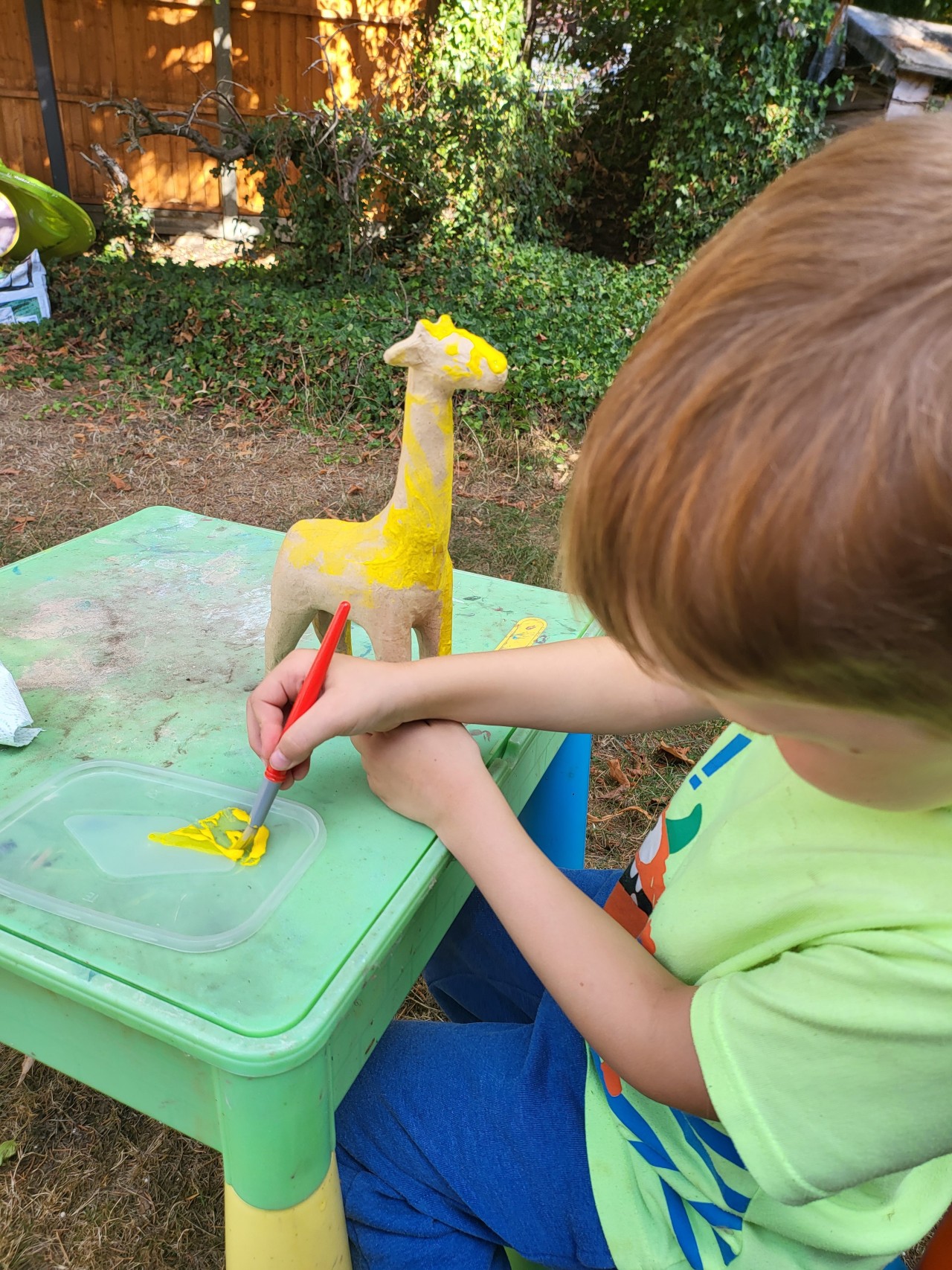 Young boy painting a cardboard giraffe yellow in the garden