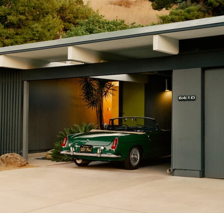 Ein Foto zeigt das Äußere eines einstöckigen Hauses in der Wüste mit einem überdachten Parkplatz, auf dem ein Oldtimer-Cabrio-Sportwagen steht.