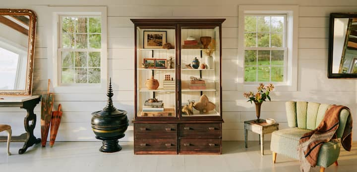 A photo shows a large white living room with a collection of antique furniture, including a large wood and glass display case full of an assortment of vintage valuables.