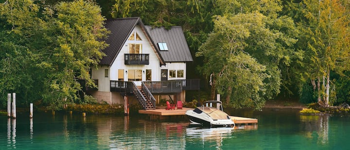 A photo shows a boat, covered and docked in front of a 2-story home on the edge of a lake.