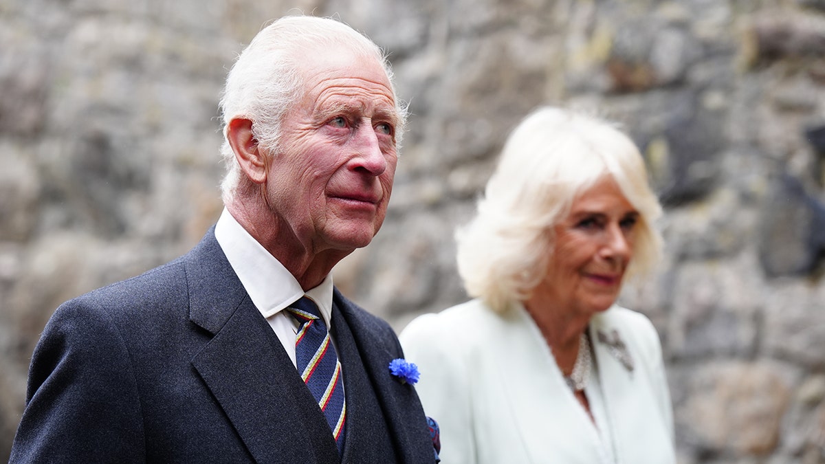 King Charles in a dark suit and striped tie walking next to Queen Camilla in a white suit.