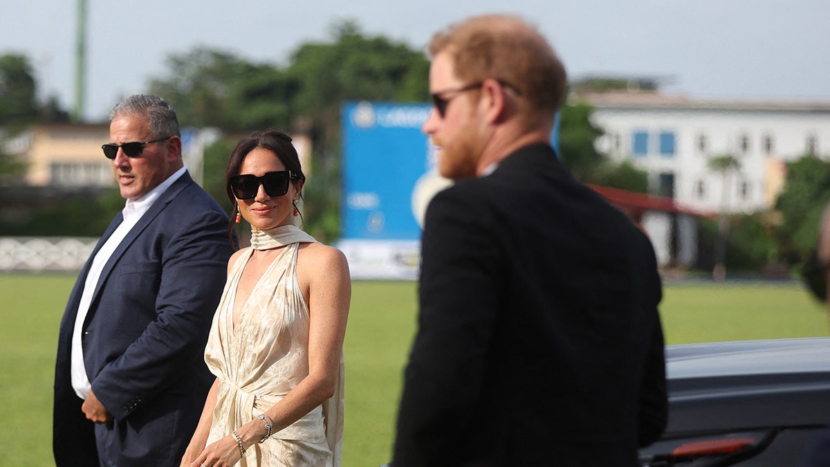 Meghan Markle in a beige dress and sunglasses, looking at Prince Harry in a black suit and sunglasses.