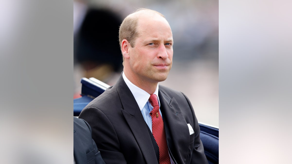 Prince William in a black suit with a red tie sitting in a carriage.
