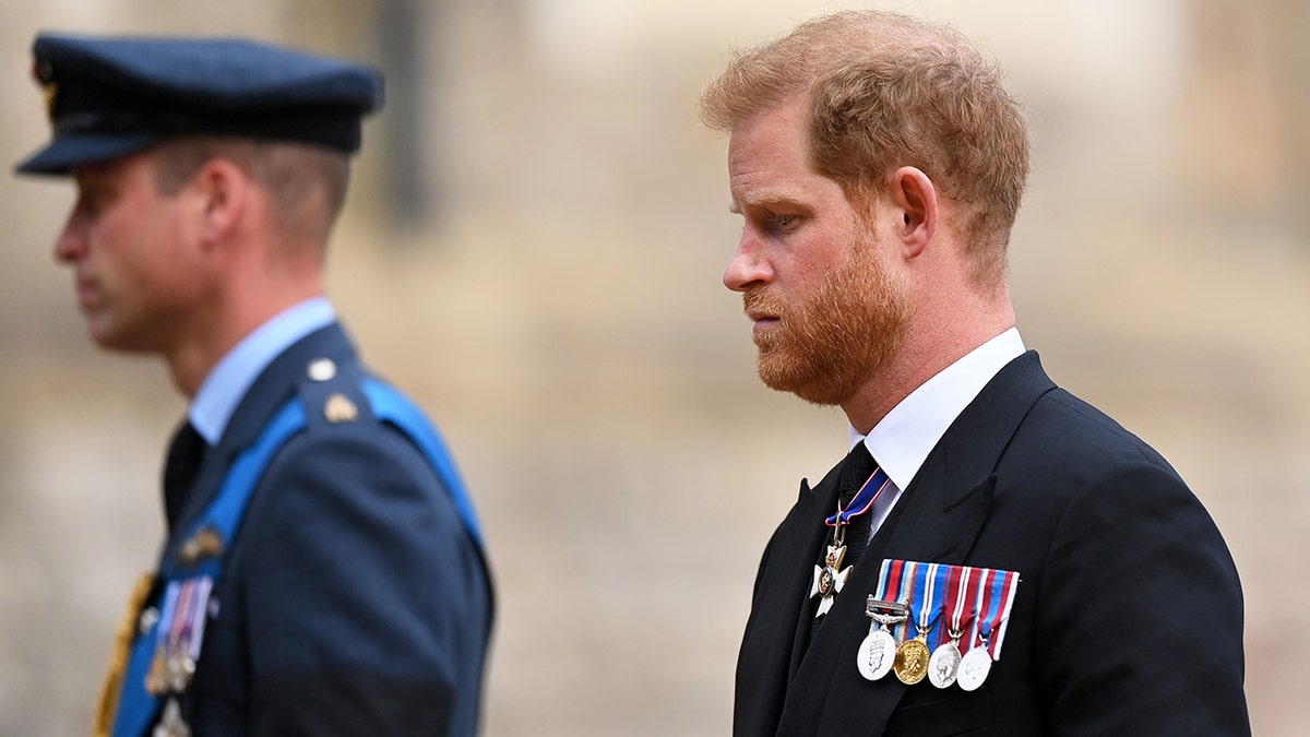 Prince Harry wearing a suit and medals looking somber as Prince William walks ahead in a blue uniform.