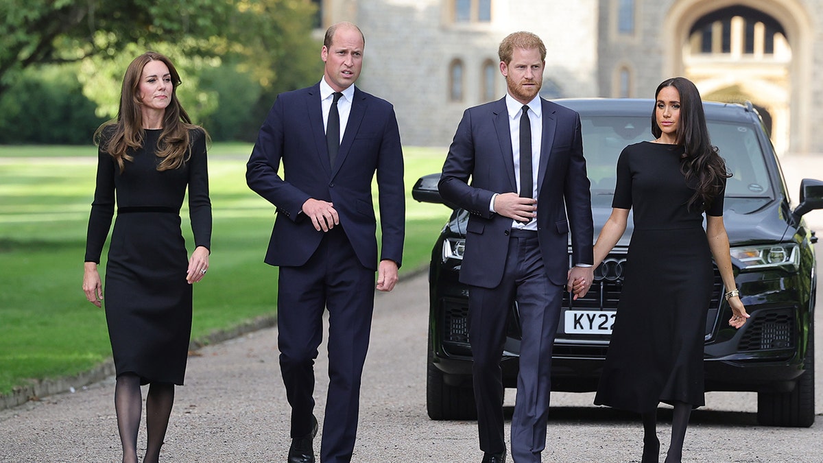 Kate Middleton, Prince William, Prince Harry and Meghan Markle walking next to each other in a line in front of a black car.