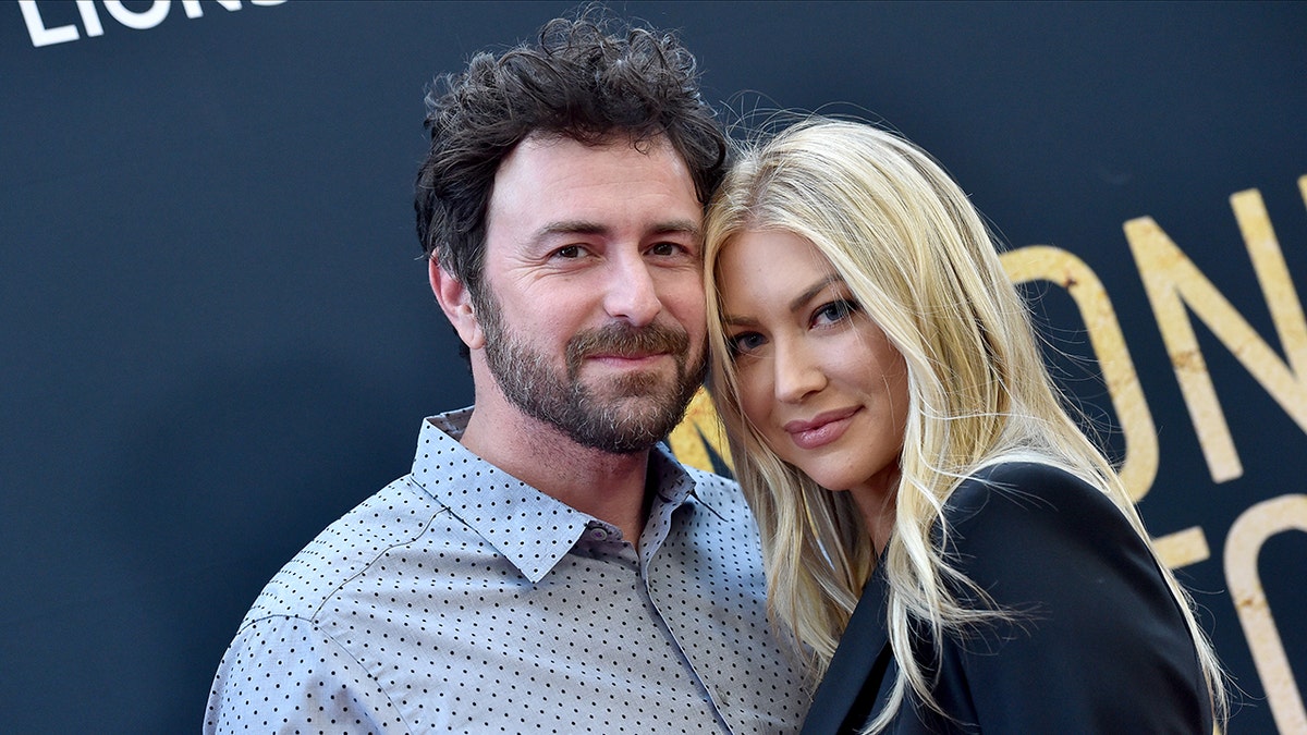 Beau Clark in a blue patterned shirt soft smiles on the carpet with wife Stassi Schroeder who leans into him