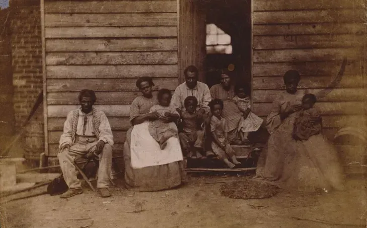 One or more families of African American slaves posed in front of a wooden house on Dr. William F. 