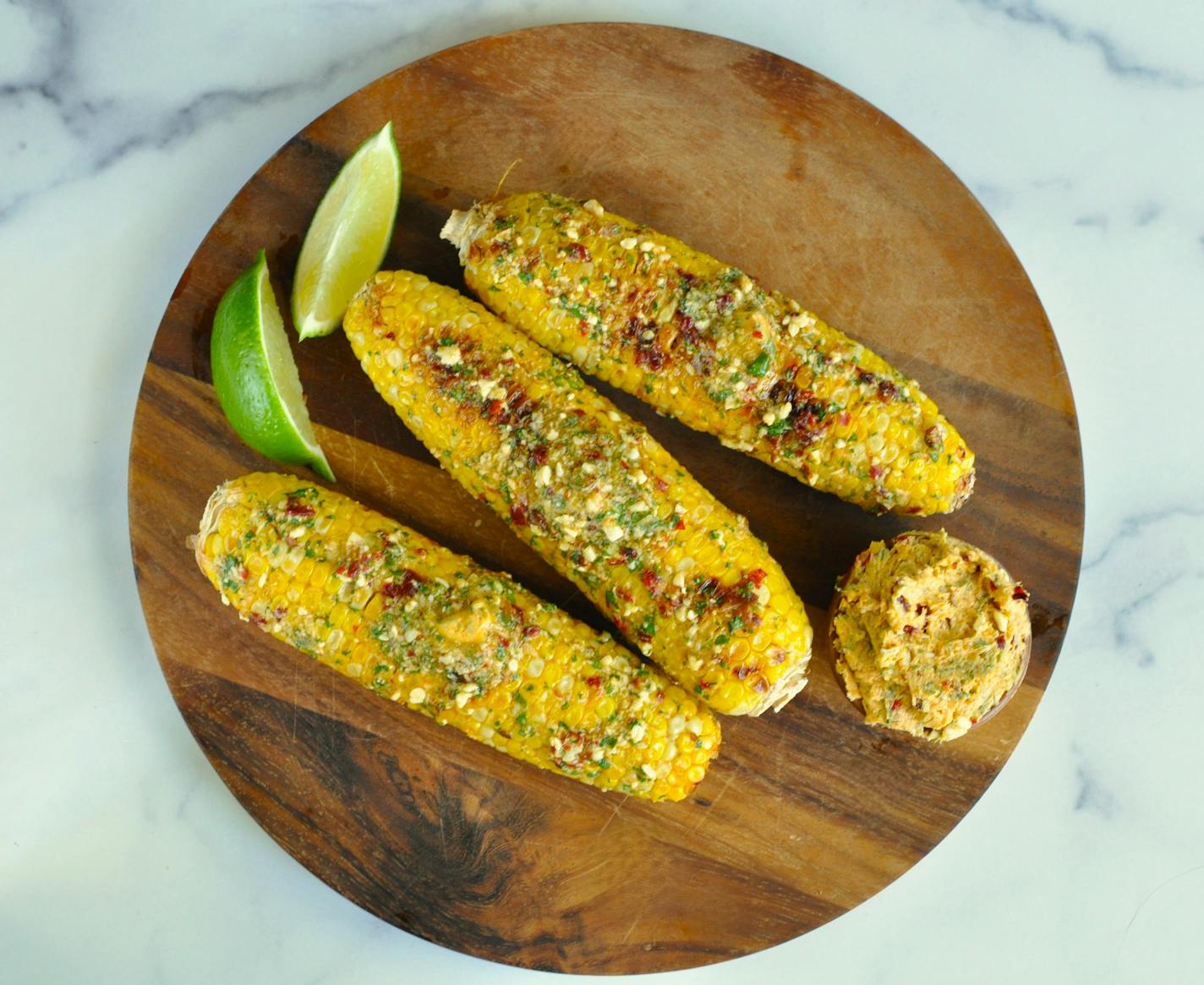 A wooden platter with three ears of grilled corn, all topped with smoky cotija butter that gets is flavor and color from a chile in adobo sauce.