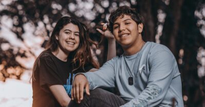 Two young teenagers look at the camera and smile. They are seated outdoors with a sunset or sunrise and the shadow of trees behind them.