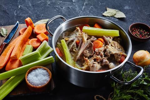 Steel pot of bone broth mixed with carrots and celery, with carrots and celery on cutting board and seasonings