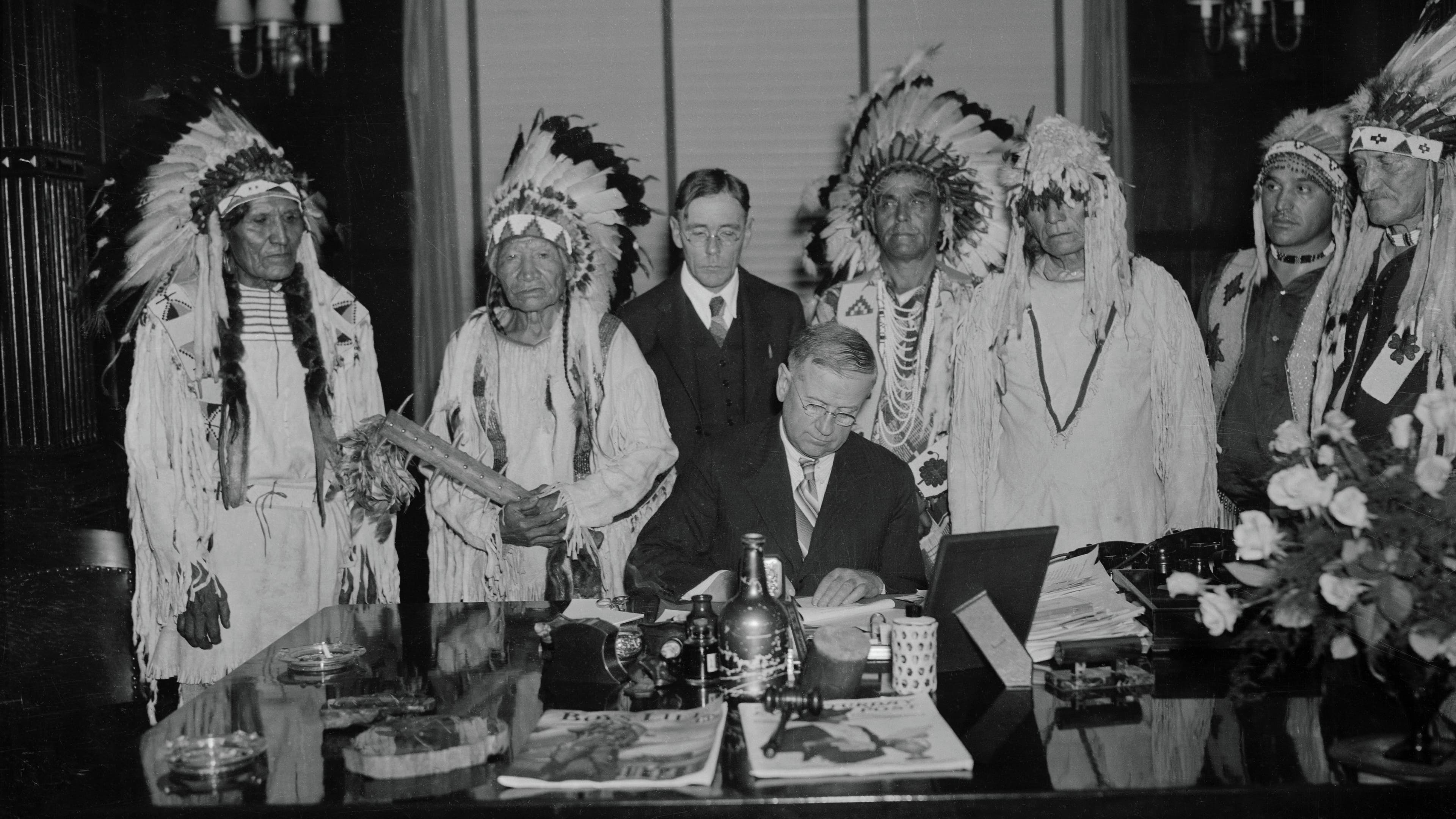 Government official in suit sitting at a desk signing a document, as a group of men in Indian clothing and headdresses stand behind him.