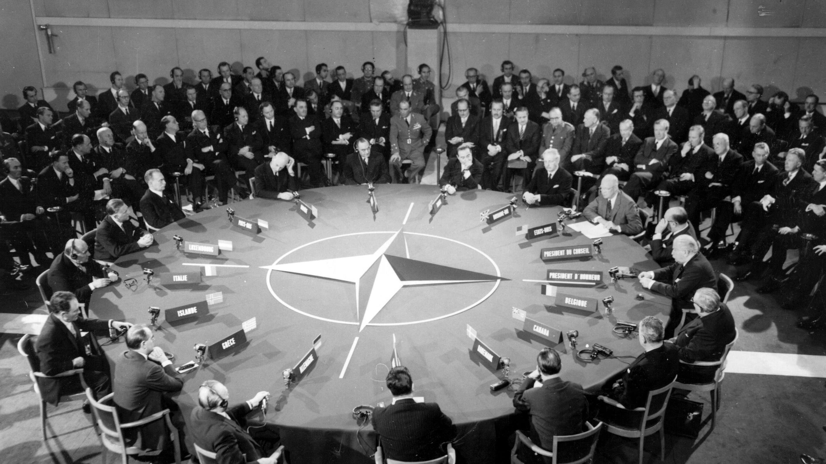 Men in suits sitting around a large round table with the NATO logo in the middle of the table.