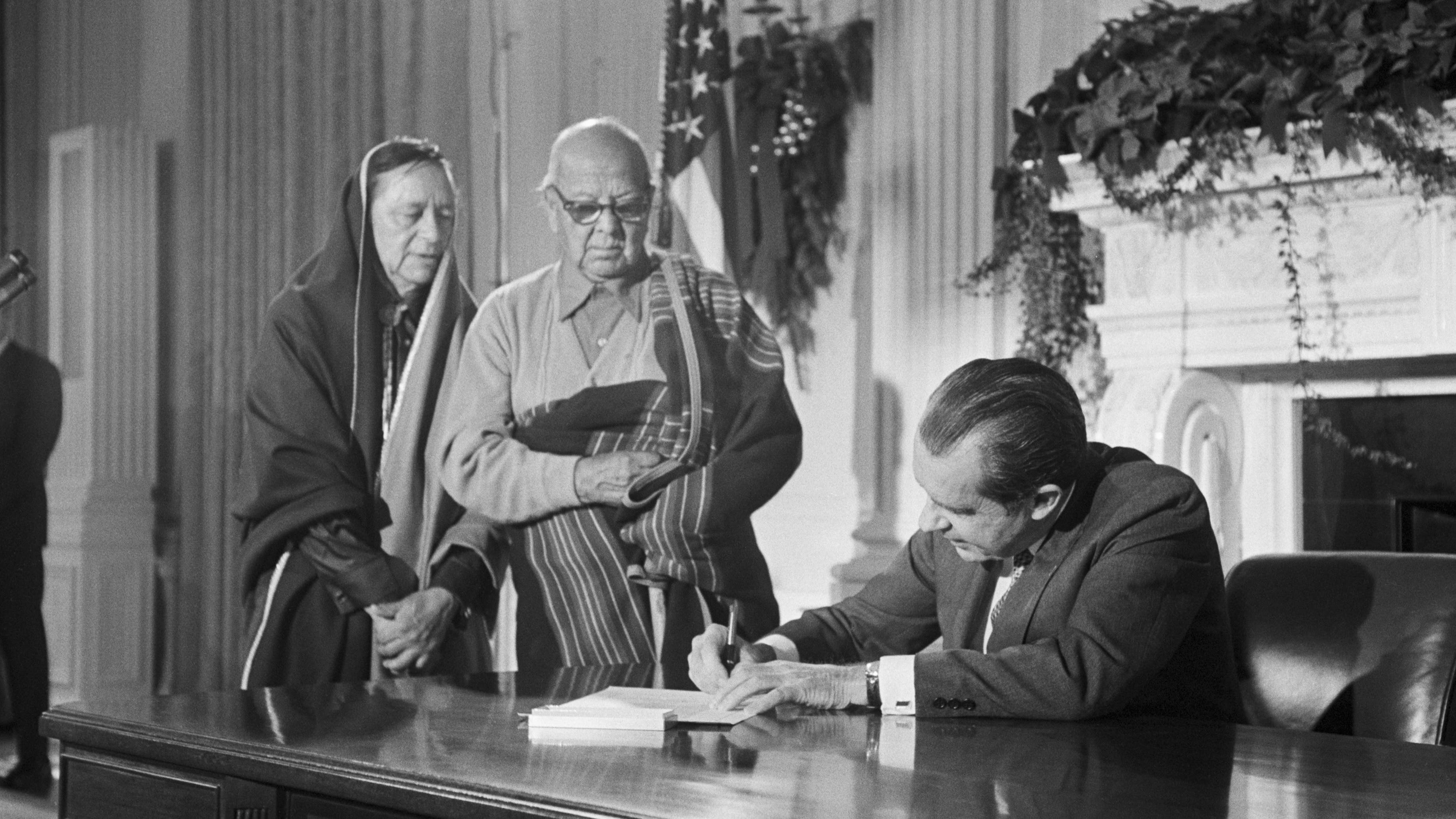 President Richard Nixon sitting at a big table signing a document while two Native American people stand to the side, looking on.