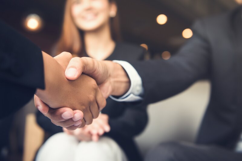 close-up of two people shaking hands while someone sits in the background smiling