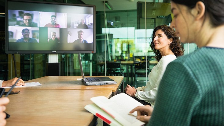 Zwei Frauen sitzen an einem Tisch, weitere Personen sind auf einem großen Bildschirm virtuell zugeschalten.