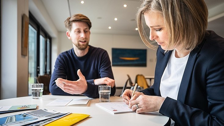 Eine Frau und ein Mann sitzen an einem Tisch auf dem Dokumente liegen, die Frau macht sich Notizen.