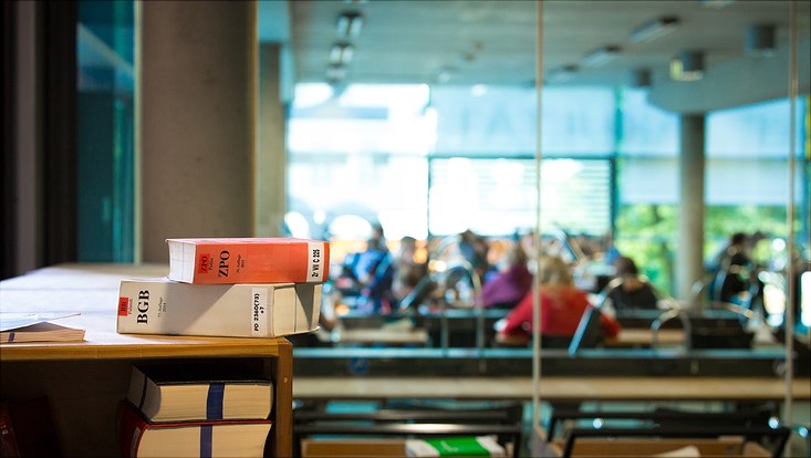 Gesetzesbücher auf einem Tisch, im Hintergrund arbeitende Studierende.