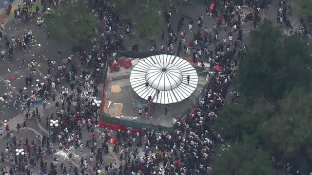 An aerial view of thousands of people crowded into Union Square. Several people are seen climbing onto the roof of a structure. 