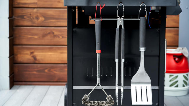 The grill utensils hang on the gas grill: a spatula, tongs, and a brush. 