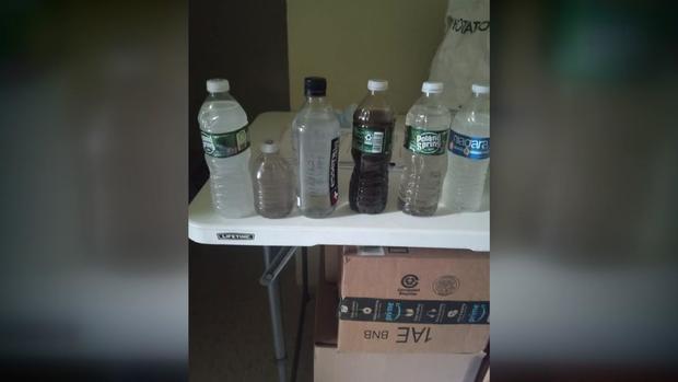 A photo of six plastic bottles of water lined up on a table. Water in one of the bottles appears black and water in at least three other bottles appears gray. 