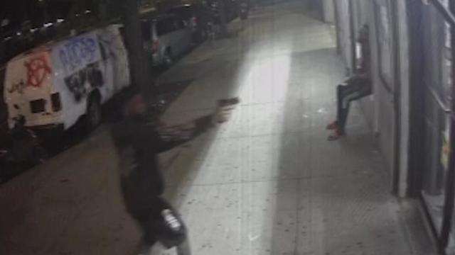 A man standing on a sidewalk points a gun at the door of a Bronx bodega while a man with a cane sits on a window ledge nearby. 