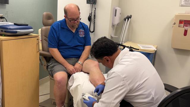 Patrick Curran sits in a chair in a hospital exam room while a doctor examines his left foot. 