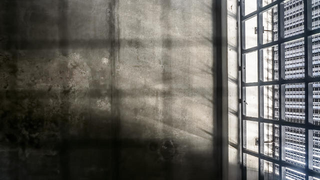 The very sober interior of a prison cell: barred windows with little light coming in and bare concrete walls 