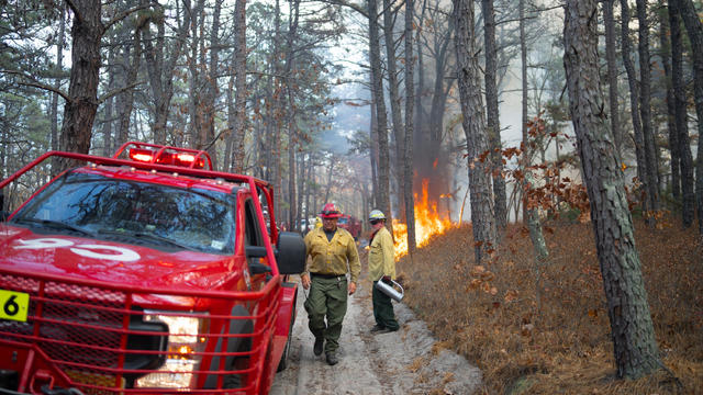 A fire truck and firefighters are seen in a forest as a fire burns in the background, hazy smoke in the background 