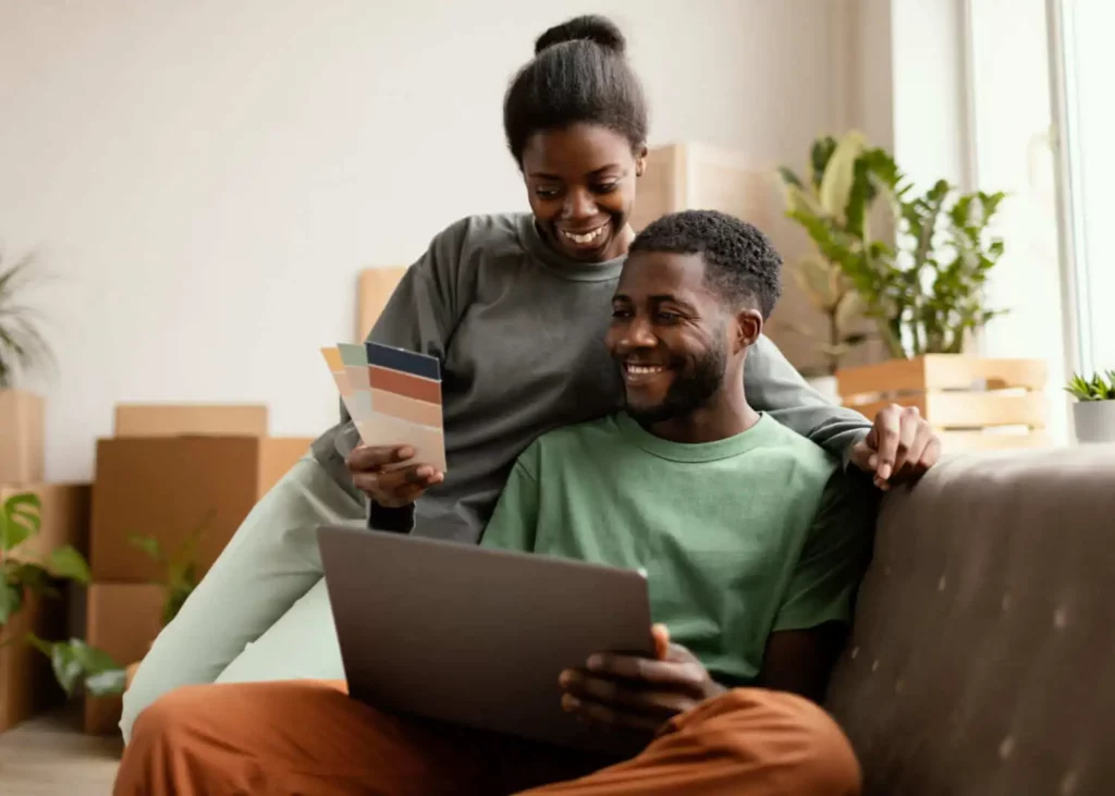 Imagem de um casal sorridente sentado com uma cartela de cores na mão em um ambiente com caixa de mudança para ilustrar matéria sobre como tirar o habite-se de um imóvel já construído 