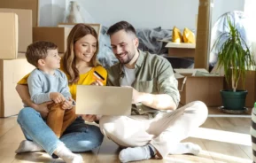 Imagem de uma família composta por um homem, uma mulher e uma criança sentados no chão de uma casa ao lado de caixas de papelão de mudança para ilustrar matéria sobre como comprar uma casa sem entrada