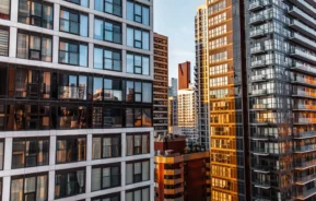 Imagem panorâmica de prédios altos de vidro e modernos com luz do pôr do sol refletida para ilustrar matéria sobre apartamento próprio