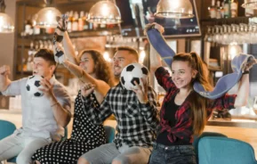 Imagem de dois homens e duas mulheres sentados no balcão de um bar torcendo com bolas de futebol na mão para ilustrar matéria sobre bar com telão