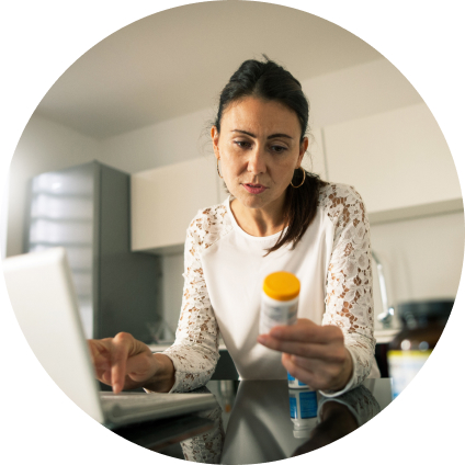 A woman reads her prescription bottle, holding it in one hand while typing on her laptop with the other